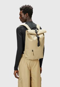 Zaino beige a rotolo con hardware nero, spallacci regolabili e tasca con zip anteriore. Realizzato in materiale resistente e impermeabile.