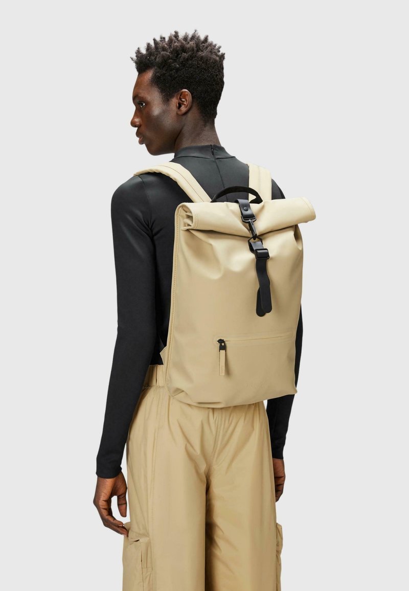 Mochila roll-top beige con herrajes negros, correas ajustables y un bolsillo frontal con cremallera. Fabricada con material duradero y resistente al agua.