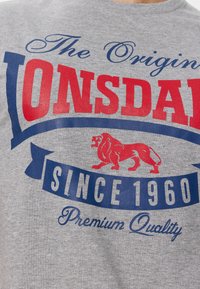 Grå bomulds t-shirt med et stort rødt og blåt grafisk design med teksten "ONSDALE," en løveillustration og teksten "SINCE 1960" i fed skrift.