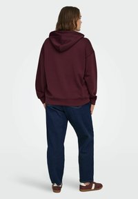 Burgundijas krāsas kapuce ar lielu kapuci, kam ir brīvs piegriezums un ribotas aproces, kombinēta ar tumši ziliem brīvi pieguļošiem džinsiem un brūnām sporta apavām.