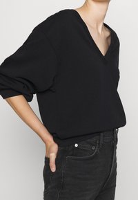 Sudadera negra con cuello en V, hombros caídos y un corte relajado. Hecha de una tela suave; presenta puños acanalados y un diseño suave y minimalista.