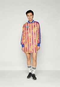 Persoană îmbrăcată într-o rochie lungă cu mânecă, colorată cu dungi, guler și manșete albastre, șosete albe Adidas și încălțăminte sport neagră, stând în picioare.