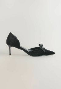 Sort ruskind, spidse kitten hæl sko med lav hæl og dekorativ sløjfe på toppen, vist i sideprofil på en neutral baggrund.