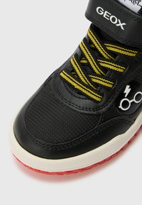 Black sneaker with yellow laces, leather and fabric upper, rubber sole, features lightning bolt and glasses graphics, red outsole accents.