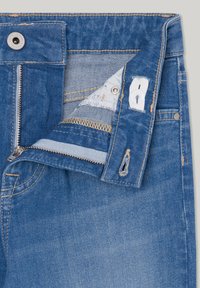 Jeans de mezclilla azul con cierre de cremallera, botón de metal y detalles de costura visibles. La parte interna de la cintura tiene una etiqueta y un forro azul claro.
