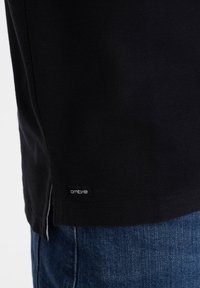 Camisa negra texturizada con una abertura lateral y una pequeña etiqueta "ombre" cerca de la parte inferior, combinada con jeans de denim azul.