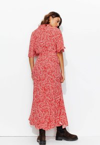 Robe maxi à motif paisley rouge avec manches courtes bouffantes, taille cintrée et ourlet froncé. Portée avec des bottines à talons marron foncé.