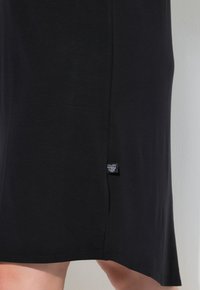 Vestido negro de longitud a la rodilla, confeccionado en una tela suave con un corte recto, que presenta una pequeña etiqueta de marca en el lado izquierdo cerca del dobladillo.