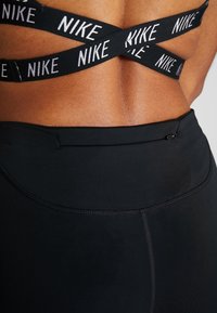 Černá sportovní podprsenka Nike s křížovým zádovým designem a bílými logy, doplněná o černé vysoce pásové sportovní legíny s malou zipovou kapsou na zadní straně.