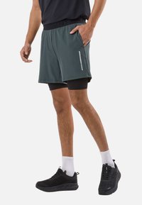 Man wearing dark green layered athletic shorts, black compression shorts underneath, white socks, and black running shoes.