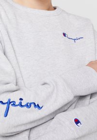 Sweatshirt gris en mélange de coton avec le logo "Champion" brodé en bleu sur la poitrine et un badge signature sur la manche.