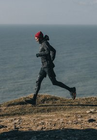 Corridore con una giacca nera leggera e leggings neri con motivo, che indossa un berretto rosso e guanti verdi, mentre corre su una collina erbosa.
