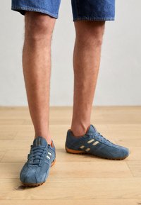 Baskets en daim bleu avec accents en mesh, rayures blanches et détail orange au talon. Semelle en caoutchouc avec une finition texturée. Portées avec un short en jean.