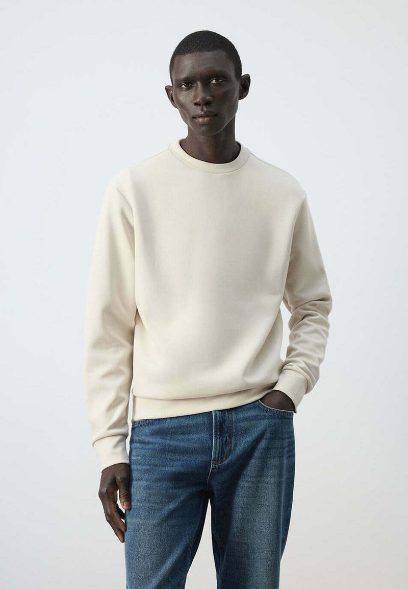 Jeune homme aux cheveux courts portant un sweat-shirt uni blanc cassé et un jean bleu, debout avec une main dans la poche devant un fond clair.
