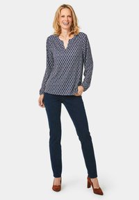 Navybluse mit weißem geometrischem Muster, V-Ausschnitt, langen Ärmeln; kombiniert mit dunkelblauen Jeans und braunen Wildleder-Highheels.