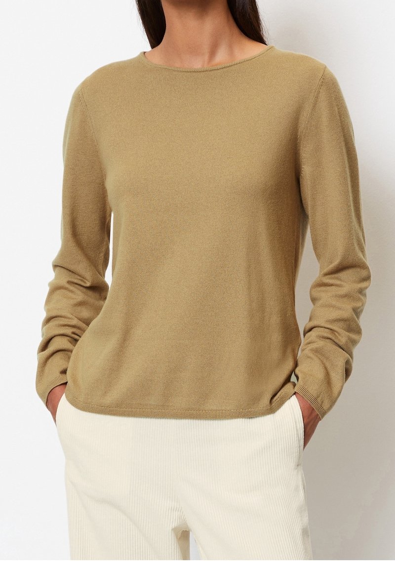Femme portant un pull en tricot beige à manches longues et un pantalon en velours côtelé écru, les mains dans les poches, sur un fond uni.