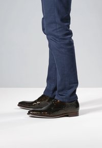 Schwarze, strukturierte Lederschuhe mit spitzer Zehenpartie, kombiniert mit marineblauen, eng anliegenden Hosen. Die Schuhe verfügen über ein glänzendes Finish und eine glatte Sohle.