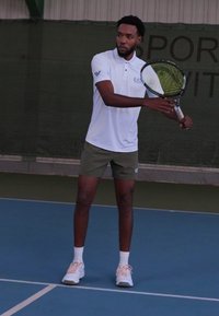 Weißes Poloshirt mit Logo, olivgrüne Shorts und weiße Sportschuhe. Die Person hält einen Tennisschläger und bereitet sich darauf vor, auf einem blauen Platz zu servieren.