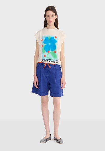 Žena stojaca, oblečená v bielom tielku bez rukávov s modrým kvetinovým vzorom a textom, modrých šortkách dĺžky po kolená s oranžovým šnúrkou a strieborných balerínach.