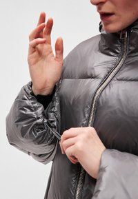 A szürke pufi dzseki magas gallérral és elöl cipzárral rendelkezik. Állítható mandzsetta húzózsinórral. Simaságával és fényes felületével, valamint steppelt részeivel tűnik ki.