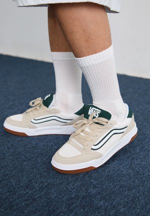 Beżowe sneakersy z zielonymi akcentami, białymi podeszwami i sznurówkami. Charakteryzują się teksturowanym materiałem, skórzanymi wstawkami oraz dyskretnym logo na języku.