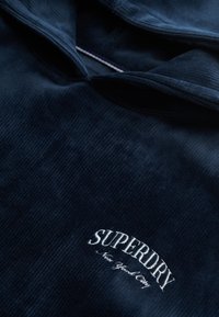Sudadera de pana en color marinero con un logo en relieve "SUPERDRY" en blanco. Presenta una textura suave y una capucha con cordón.