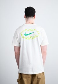 Hvid bomulds T-shirt med en grøn-blad omgivelse og et blåt Nike-logo på ryggen. Korte ærmer og standard pasform.