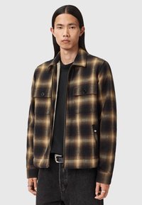 Giacca svasata a quadri nera e marrone con zip frontale, due tasche sul petto e una morbida fodera interna. Indossata sopra una maglietta nera e jeans scuri.