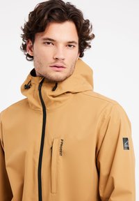 Chaqueta impermeable color beige con cuello alto, capucha ajustable, cremallera frontal y bolsillo en el pecho. Presenta cremalleras y detalles del logo en negro.