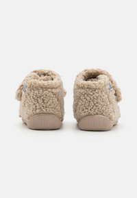 Chaussons en beige moelleux avec un extérieur texturé et duveteux, un dos arrondi et des semelles en caoutchouc. Présentent un tissu doux et un design simple.