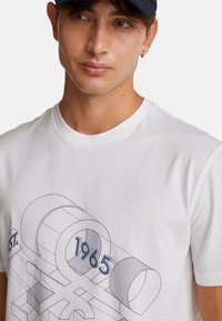 Bílá bavlněná tričko s modrým geometrickým grafickým designem se vzorem „1965“ a linkami, kulatým výstřihem, krátkými rukávy a hladkou strukturou.