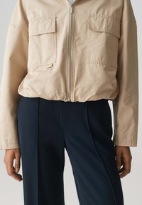 Beige cropped jack met een hoge kraag, twee voorzakken en een zilveren rits, gecombineerd met donkerblauwe broeken met een gladde textuur.