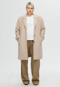 Manteau en laine beige avec un col cranté et deux boutons, associé à une chemise blanche et un pantalon marron taille haute. Texture lisse.