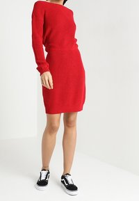 Vestido de punto rojo con mangas largas y corte relajado, que presenta una textura acanalada y un lazo en la cintura, combinado con zapatillas negras y blancas.