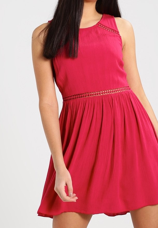 Femme portant une robe rouge sans manches avec un détail en dentelle à la taille et à l'encolure, debout devant un fond clair uni.