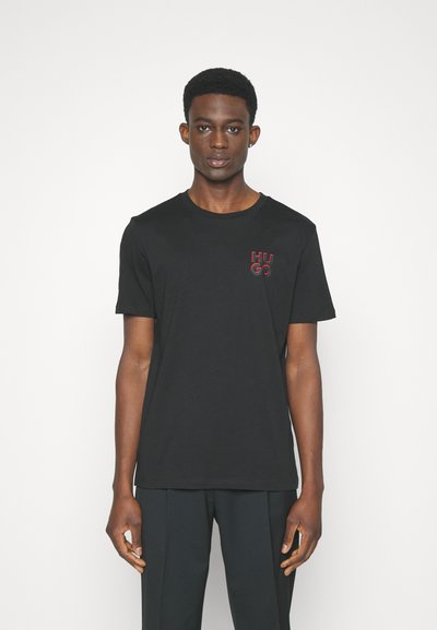 T-shirt en coton noir avec un col rond, des manches courtes et un logo rouge "HUGO" sur la poitrine gauche. Texture lisse, coupe standard.