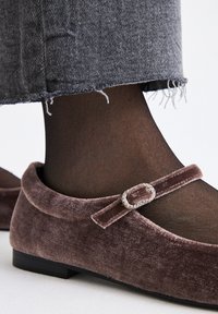 Zapatos planos de terciopelo en color malva, con punta redondeada, una hebilla decorativa y medias negras ligeras; combinados con unos vaqueros grises deshilachados.