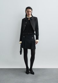 Juodas trumpas švarkas su auksinėmis detalėmis, tekstūrinis juodas megztinis, plisėta juoda sijonas, juodos pėdkelnės ir juodos aukštakulnės bateliai. Minimalistinis dizainas.