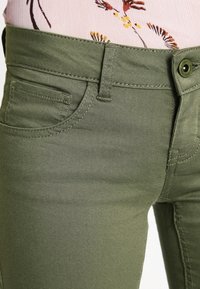 Jeans de mezclilla verde oliva con corte slim. Presentan bolsillos delanteros, cierre de botón y detalles de costura a lo largo de las costuras.