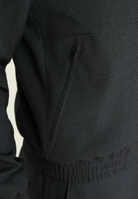 Ciemna kurtka z wzorem, z kieszenią na zamek, elastycznym pasem i fakturowanym materiałem, pokazana z boku.