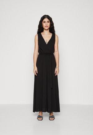 Robe maxi noire sans manches avec un décolleté en V, taille cintrée et jupe fluide. Portée avec des sandales à glissière grises. Tissu lisse, design minimaliste.