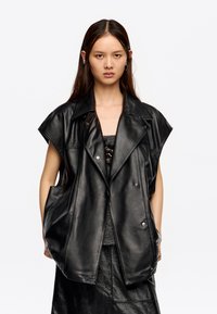 Melns ādas bezrūpju vests ar platām apkaklēm, īsām piedurknēm un pogu aizdari. Iezīmē priekšējās kabatas un gludu, spīdīgu tekstūru.