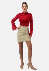 Top corto in pizzo rosso con maniche a campana lunghe, abbinato a una gonna a tubino beige con dettaglio dei bottoni. Modella indossa tacchi nudi.