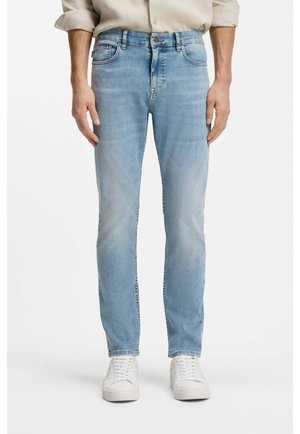 Mężczyzna noszący jasnoniebieskie dopasowane jeansy, beżową koszulę z długim rękawem i podwiniętymi mankietami oraz białe trampki, stojący na białym tle.