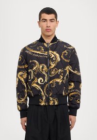 Svart kviltad bomberjacka med gyllene snurrande mönster och "Versace Jeans Couture"-text, med svart ribbad krage och mudd.