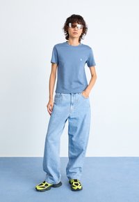 Person som går i blå t-skjorte, lyseblå vide jeans, neongule og svarte joggesko, og reflekterende solbriller som står på blått gulv.