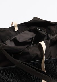 Czarna torba typu tote z zewnętrzną siateczkową częścią i materiałową podszewką. Posiada beżowe paski i górne otwarcie zapewniające dostęp do przestronnego wnętrza.