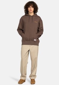 Un sweat à capuche brun avec poche avant, cordon de serrage et le logo "Element" en orange. Associé à un pantalon beige et des chaussures brunes.