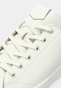 Bílé kožené tenisky s hladkým povrchem, vystouplé bílé šňůrky, kulaté otvory na šňůrky a geometrická nášivka na jazyku.