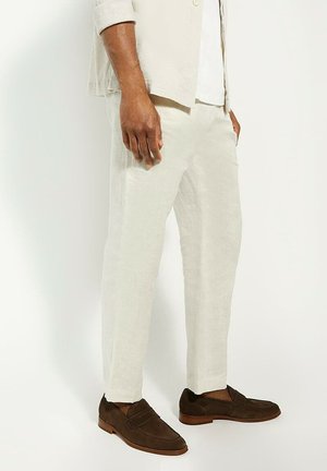 Light beige linen pants with a relaxed fit, tapered leg, and two side pockets. Paired with dark brown suede loafers.